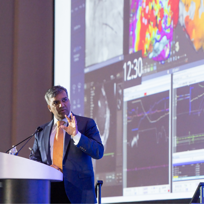 A physician presents at a podium during Heart Rhythm 2026, gesturing toward a large screen displaying cardiac mapping images and electrophysiology data.
